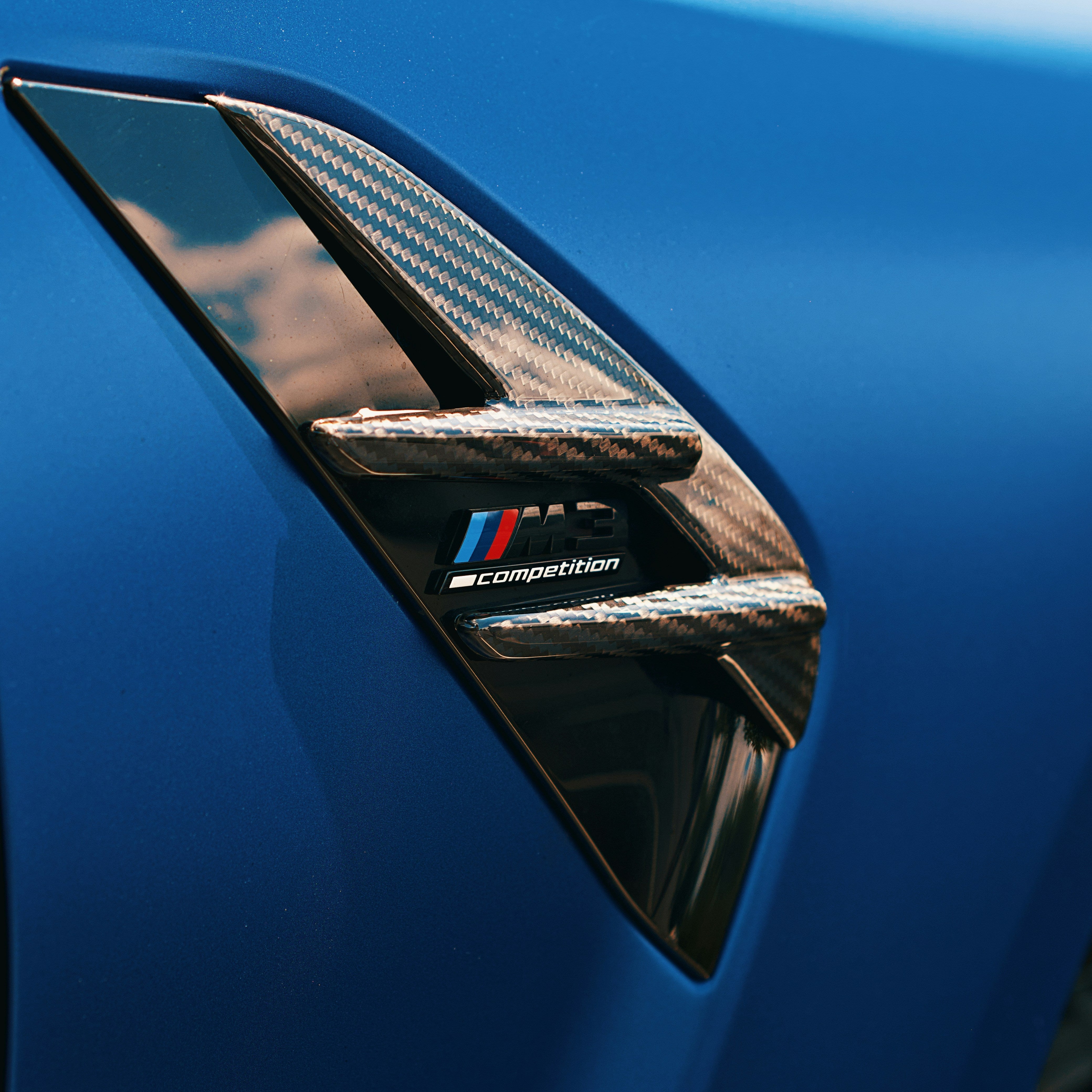 Close-up of a blue car with a carbon fiber air vent and 'M' logo.